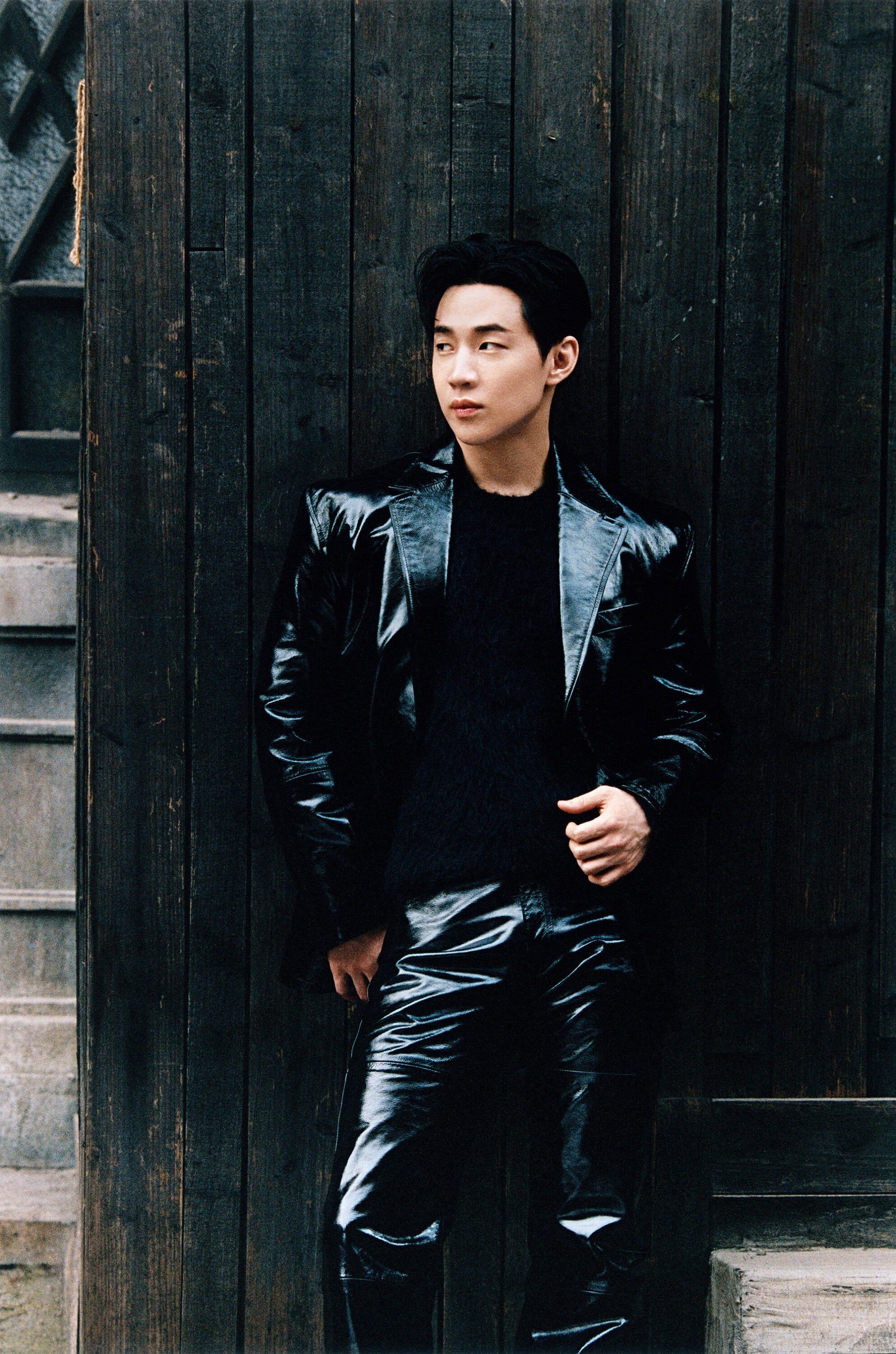 Model Henry Lau in black patent leather jacket and trousers posing against dark wooden wall for Ami Fall 2024 campaign