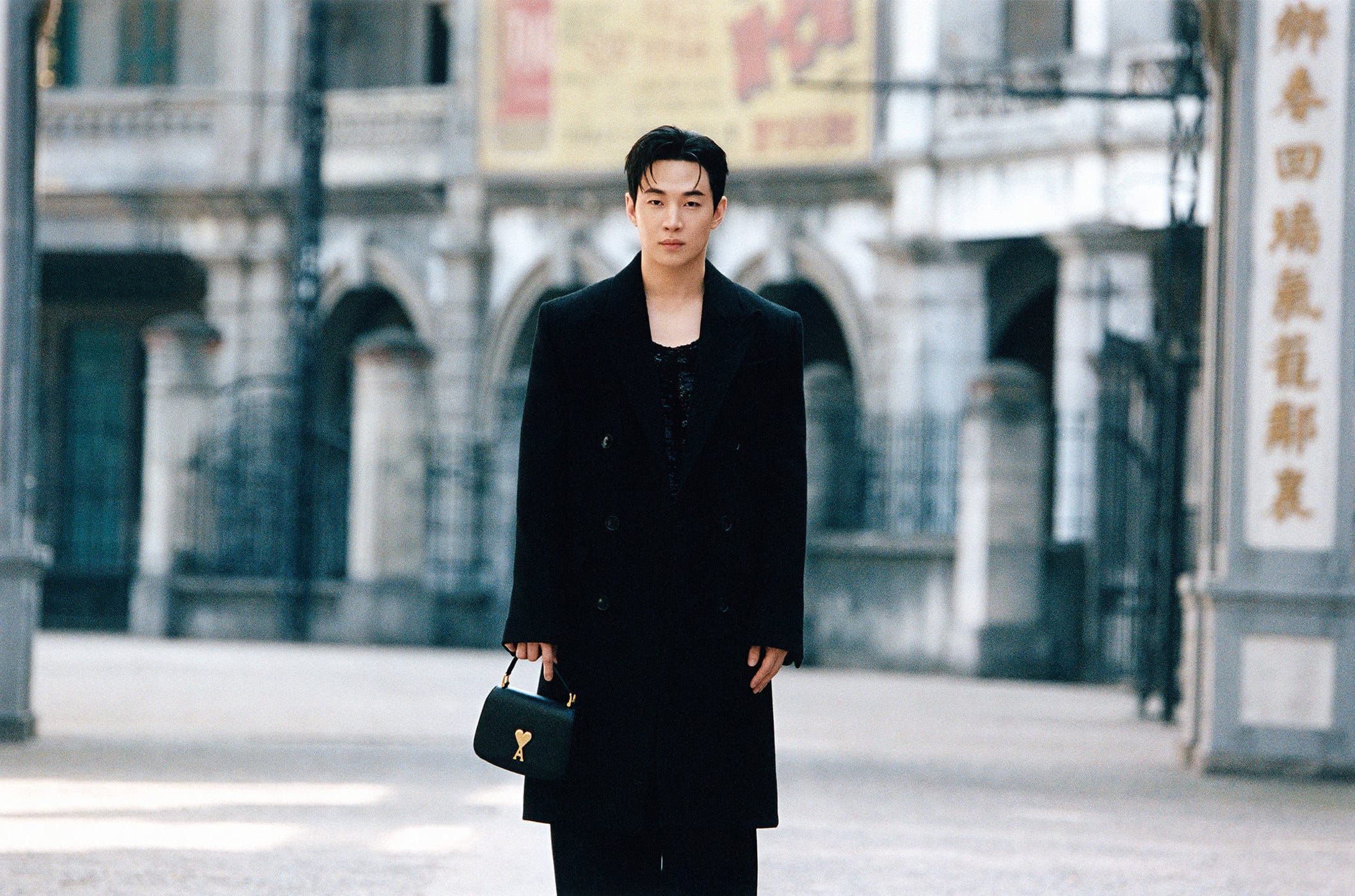 Model Henry Lau standing outdoors wearing a black coat and holding a black handbag for Ami Fall 2024 ad campaign