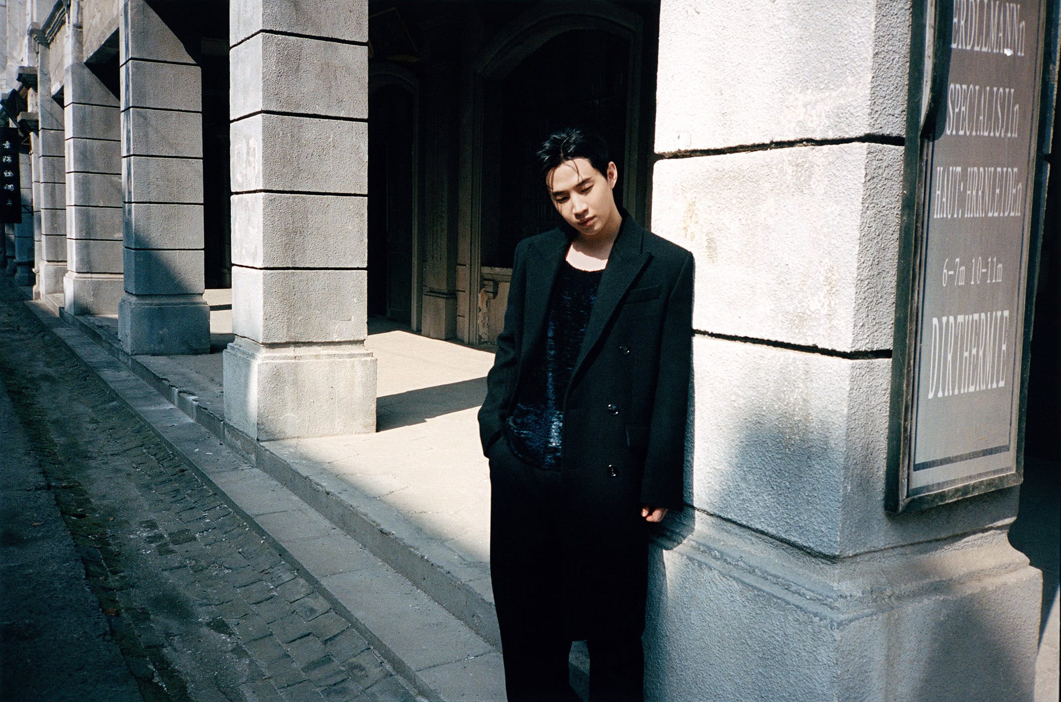 Model leaning against a stone building pillar wearing a dark tailored coat and black trousers