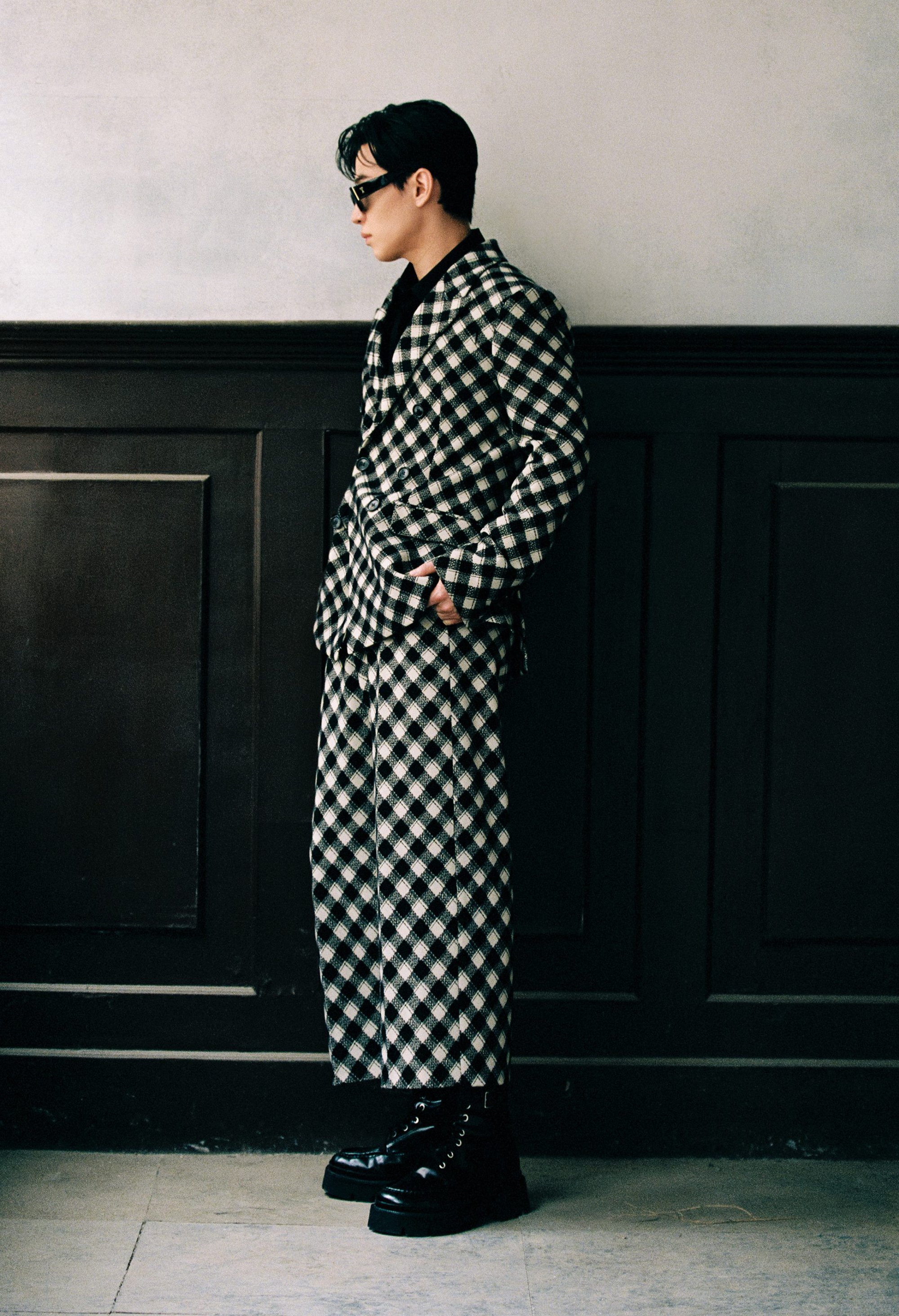 Model wearing a black and white checkered suit and sunglasses standing against a dark paneled wall