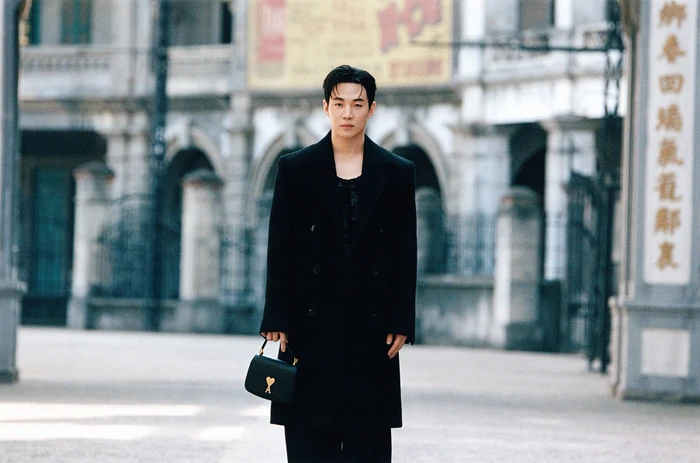 Model Henry Lau standing outdoors wearing a black coat and holding a black handbag for Ami Fall 2024 ad campaign