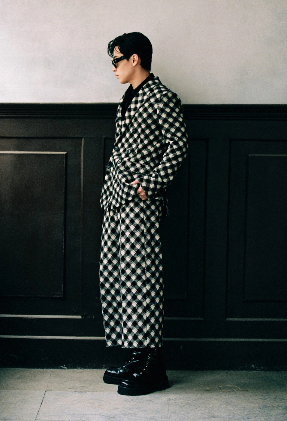 Model wearing a black and white checkered suit and sunglasses standing against a dark paneled wall