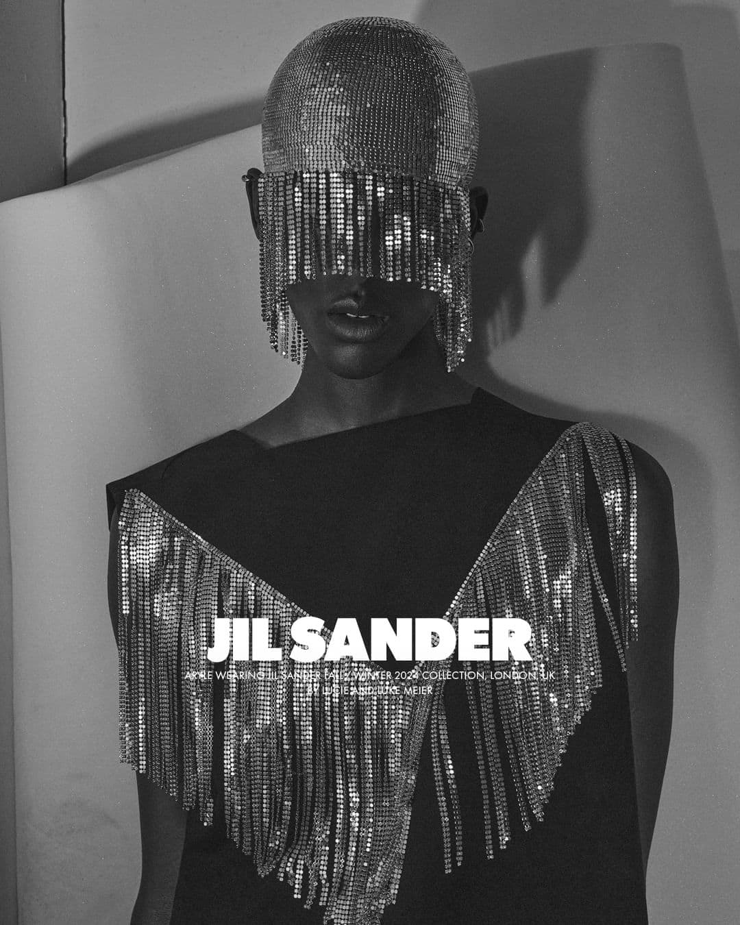 Model wearing a heavily jeweled fringe headpiece and matching crystal trim dress against a white seamless backdrop for Jil Sander.