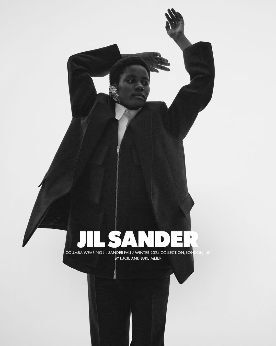 Monochrome portrait of a model in a black jacket and white collar shirt with arms raised against a plain background