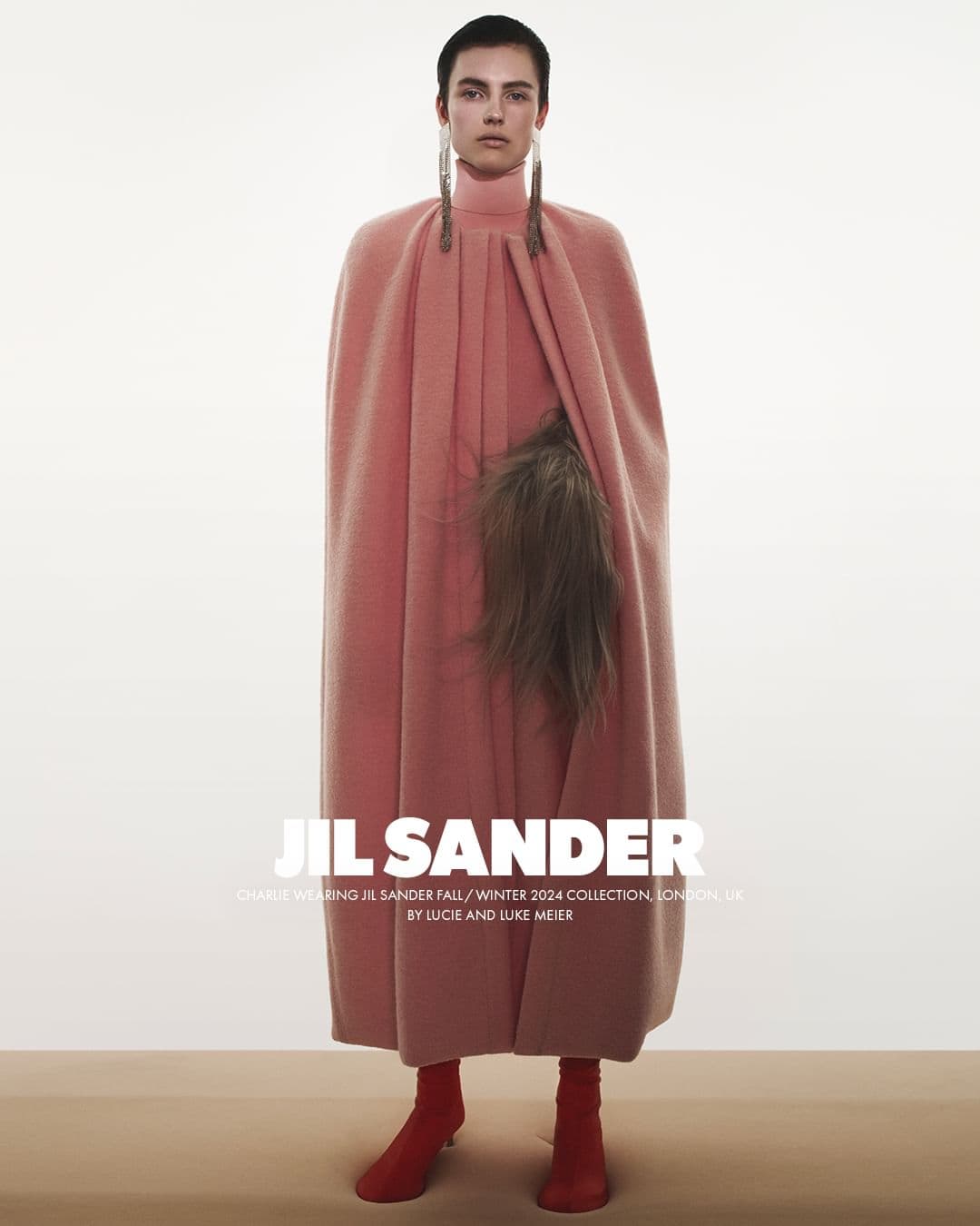 Model wearing a structured pink wool cape and matching turtleneck with long fringe earrings standing against a white backdrop.