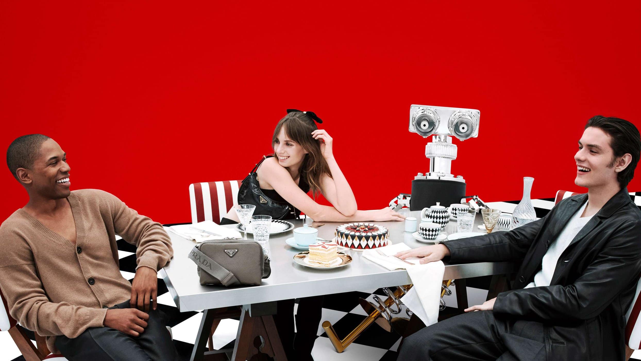 Three people seated around a metallic dining table with a robot figure against a solid red backdrop for Prada.