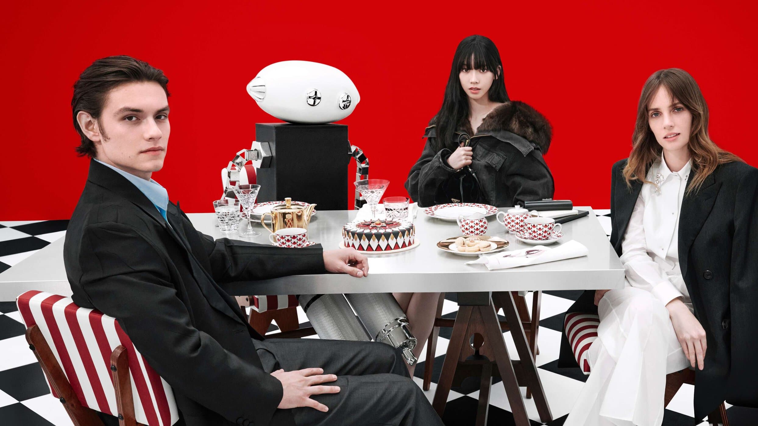 Four people seated around a metallic dining table against a solid red wall and black-and-white checkered floor for a Prada campaign