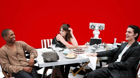 Three people seated around a metallic dining table with a robot figure against a solid red backdrop for Prada.