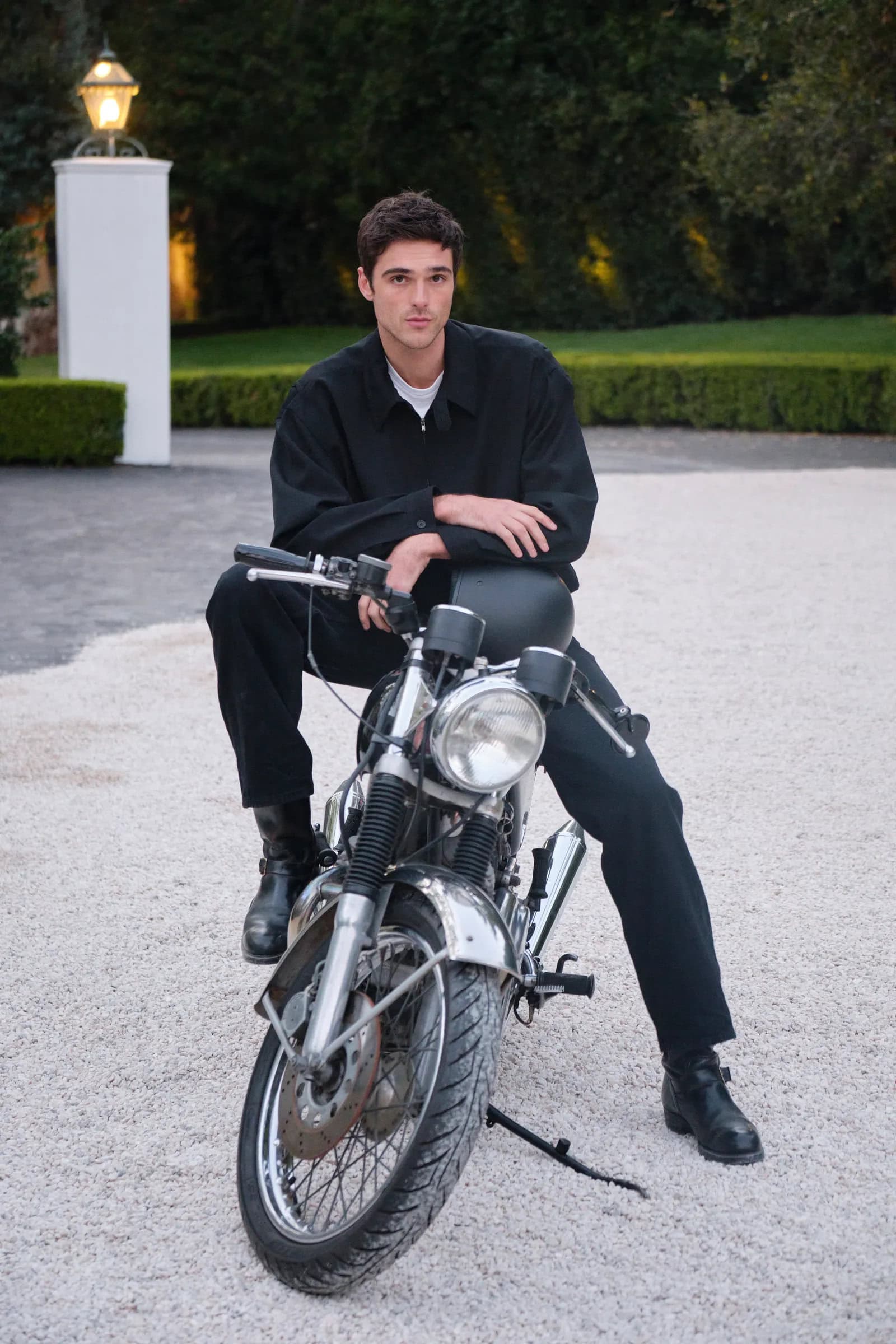 Jacob Elordi seated astride a motorcycle wearing a black shirt and trousers on gravel