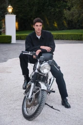 Jacob Elordi seated astride a motorcycle wearing a black shirt and trousers on gravel