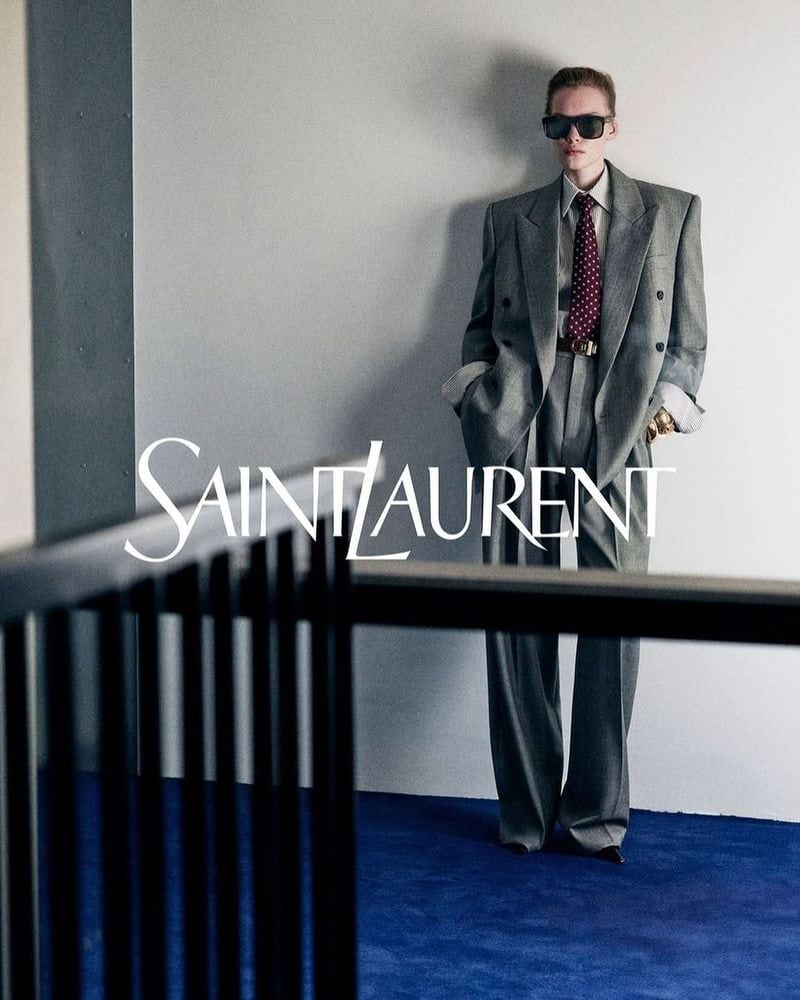 Model wearing a grey oversized suit and tie standing against a white wall above a blue carpet, framed by dark architectural railings.