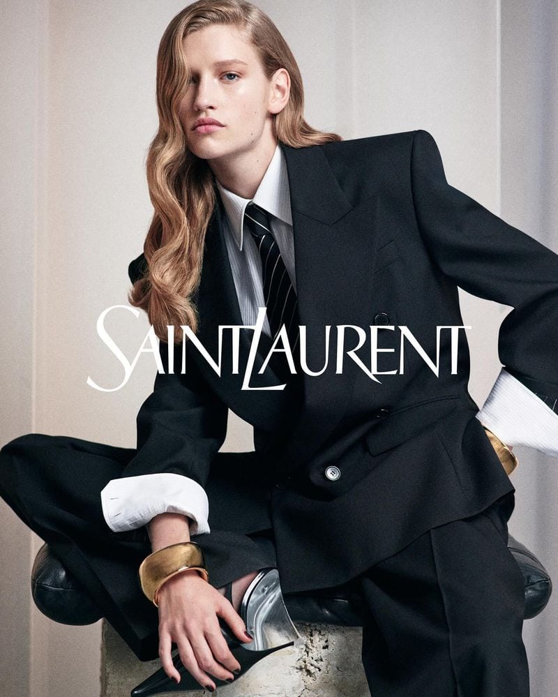 Model seated wearing a black blazer, white shirt, and tie with a large gold cuff bracelet for Saint Laurent
