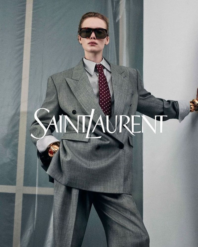 Model in grey double-breasted suit wearing sunglasses and a patterned tie against a light grey wall backdrop.