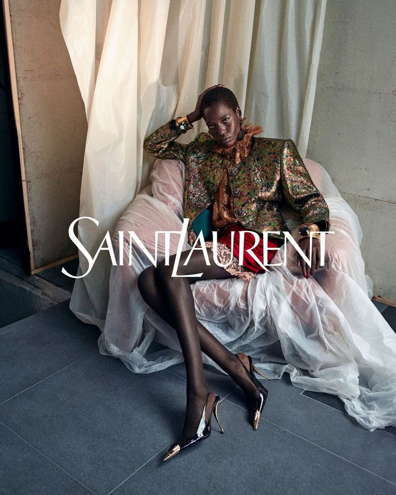 Model seated in an armchair draped in white fabric wearing a metallic patterned jacket and black heels with metallic toes