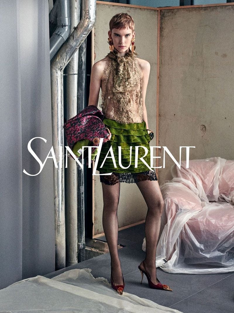 Model in green ruffled skirt and sheer halter top holding a patterned jacket against an industrial backdrop