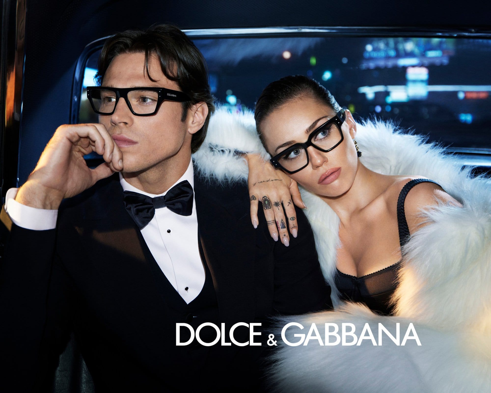 Miley Cyrus and a man wearing black eyeglasses seated inside a vehicle at night for Dolce and Gabbana eyewear campaign