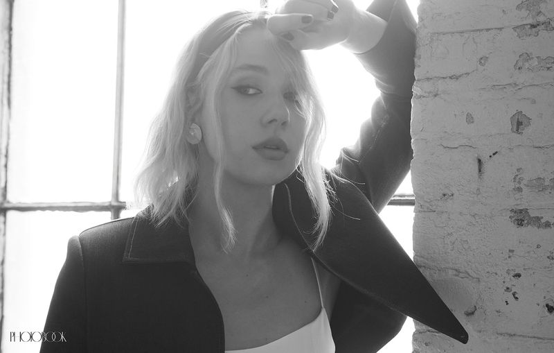 Black-and-white portrait of a blonde woman in a blazer, looking at the camera, leaning against a white brick wall in front of a window.