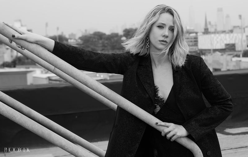 Black-and-white portrait of a blonde woman in a black blazer, posing on a rooftop with a cityscape in the background.