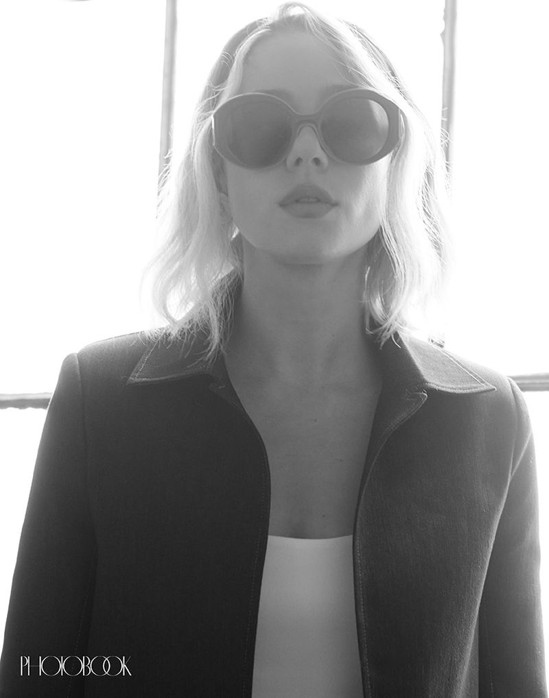 A monochrome portrait of an adult female wearing sunglasses and a black jacket, posing in front of a bright window in an indoor setting.