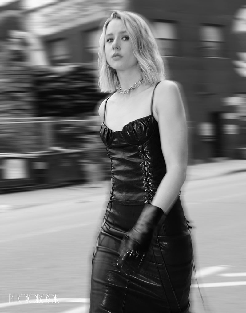 Monochrome image of a young woman in a black dress and gloves walking in an urban setting with blurred motion in the background.