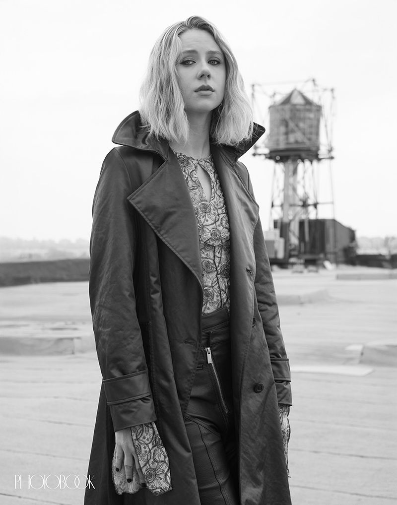 Black and white portrait of a blonde woman in a coat on a rooftop, with a vintage water tower in the distance against a bright sky.