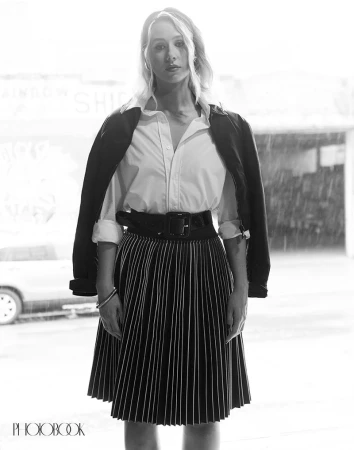 A young woman in a white shirt and striped skirt stands in front of a blurred urban storefront on a rainy day in a black and white fashion shot.