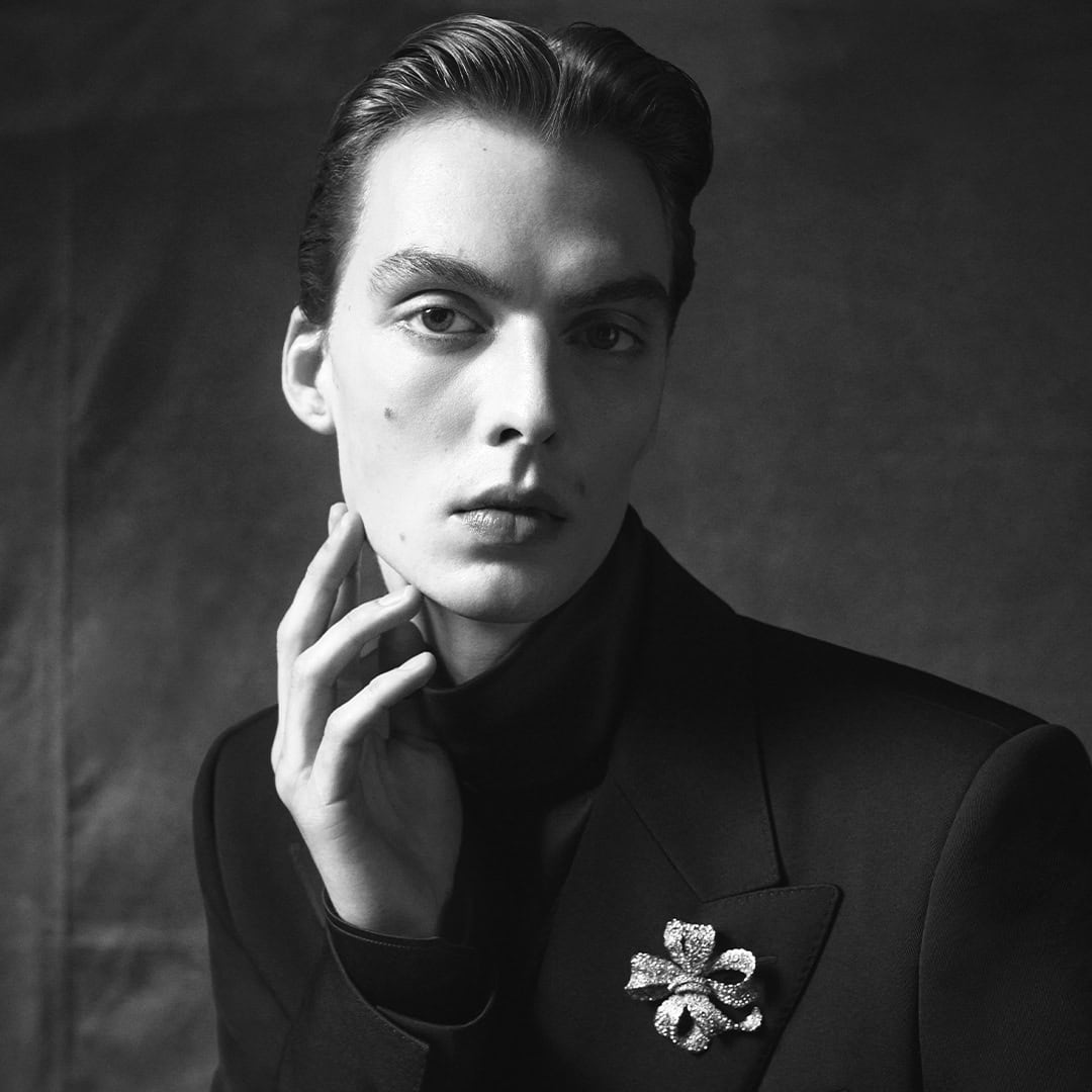 Black and white portrait of male model Leon Dame wearing a dark jacket with a jeweled brooch for Dolce & Gabbana.