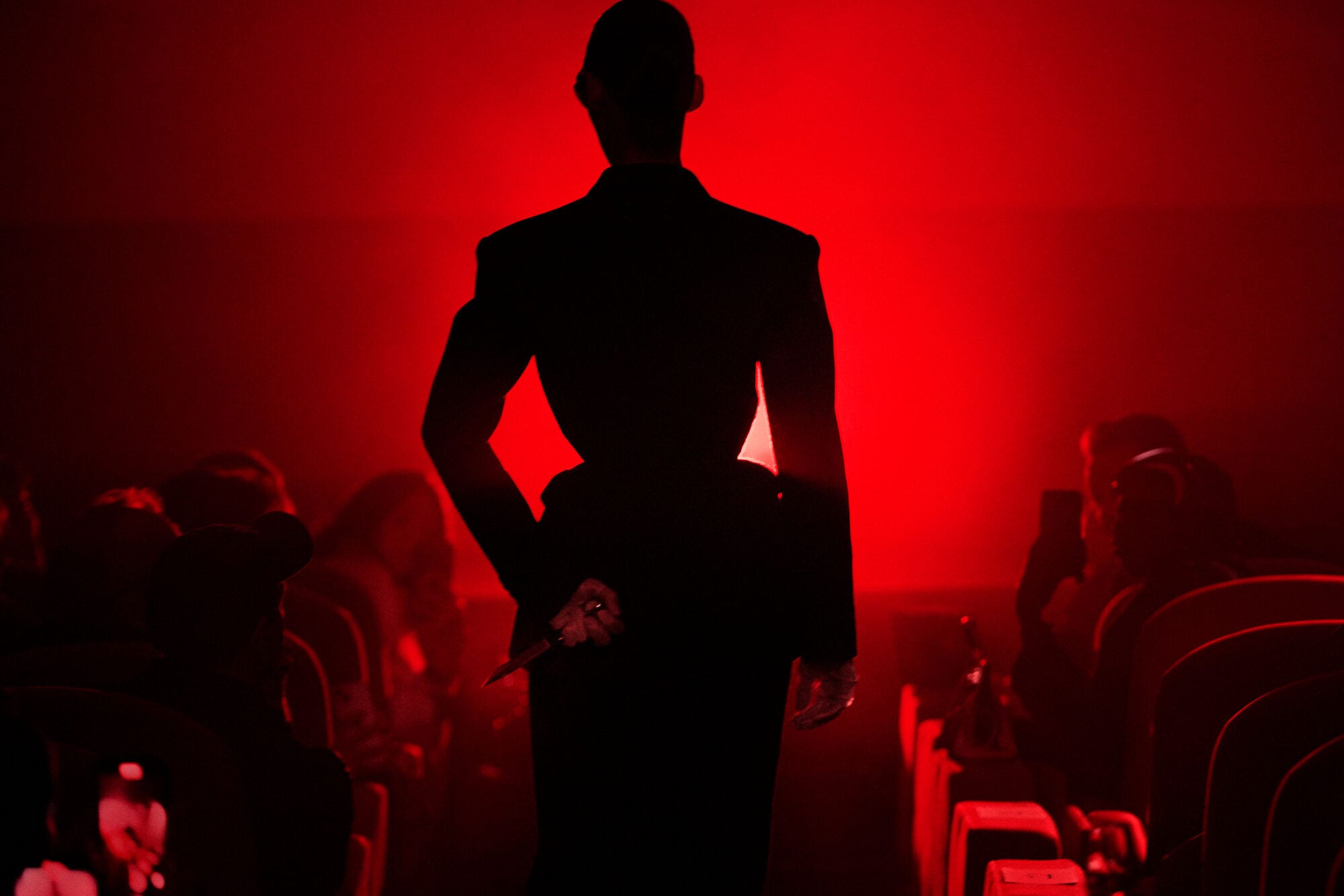 Silhouette of a person standing centrally in a dark tailored coat against an intense red backdrop in a theater setting