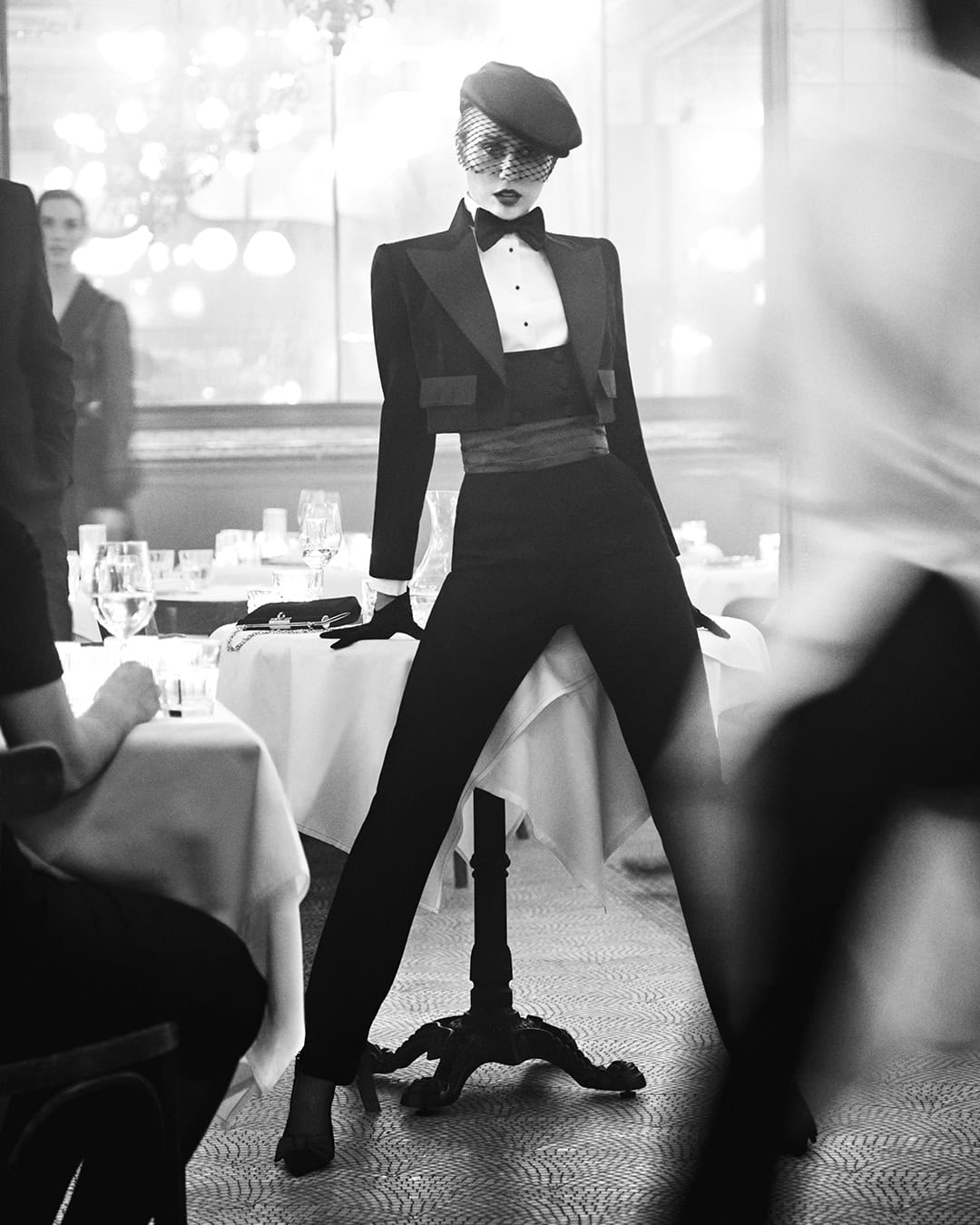 Black and white editorial photograph of a model in a tailored tuxedo leaning on a restaurant table