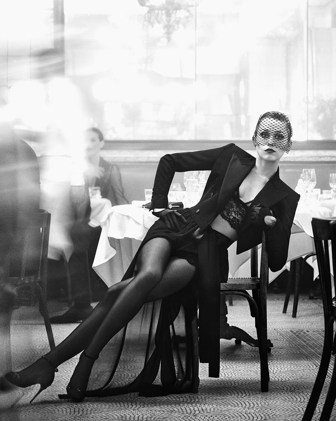 Model wearing a black blazer, lace bralette, and veil seated at a restaurant table in a monochrome campaign image