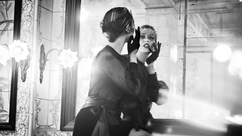 Monochrome image of a woman wearing a netted veil and black gloves adjusting her hair in a vintage mirror.
