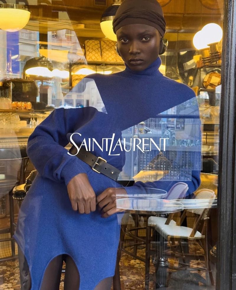 Model wearing a blue turtleneck dress and dark head wrap standing behind a glass window with Saint Laurent text