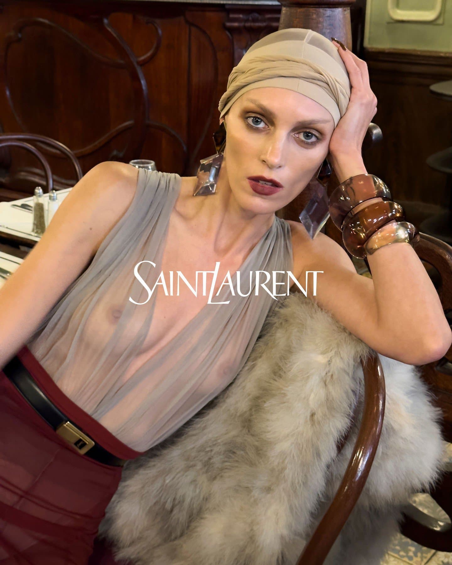Model wearing a sheer grey dress, head wrap, and chunky bracelets seated in a restaurant for Saint Laurent campaign