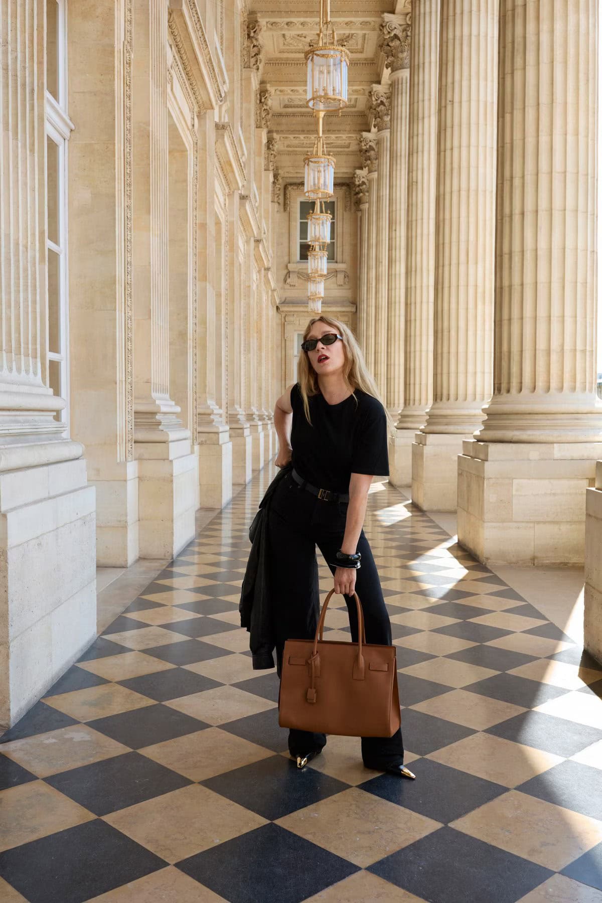 Chloe Sevigny holding a brown leather tote bag standing along a colonnade with a checkerboard floor for Saint Laurent.