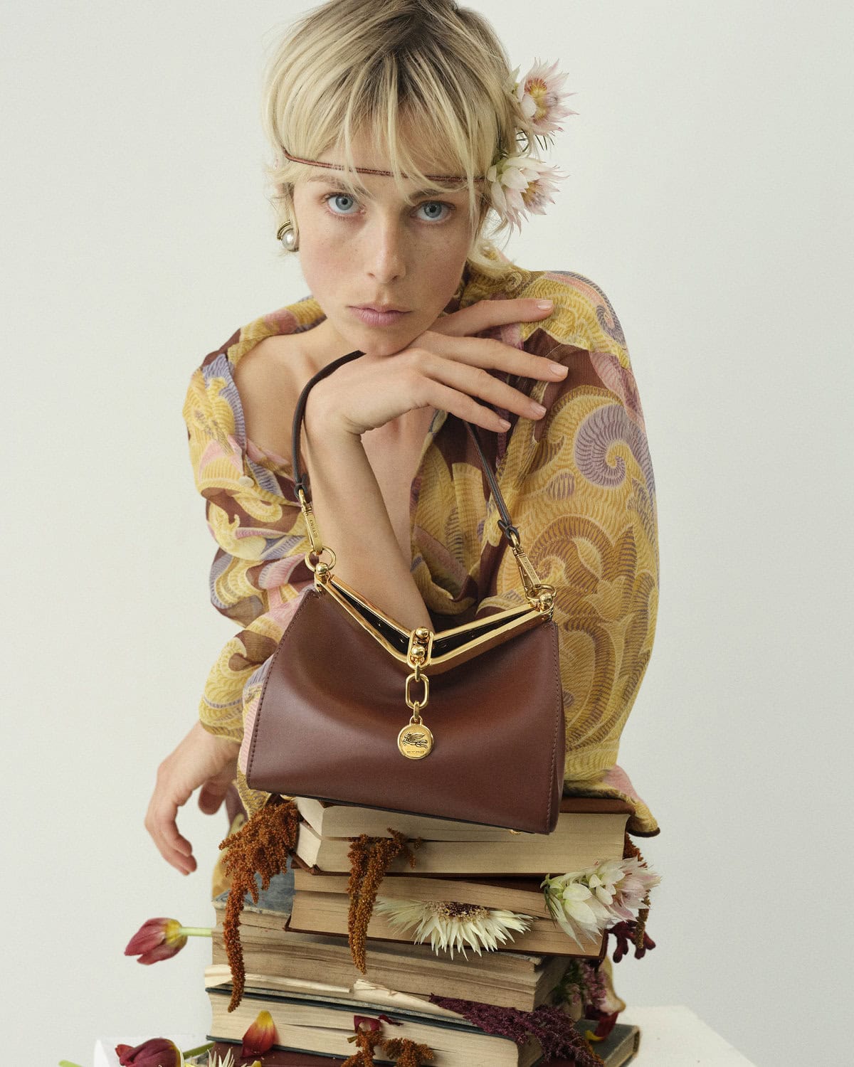 Model Edie Campbell holding an Etro Vela brown leather bag resting on a stack of books for the 2024 campaign.
