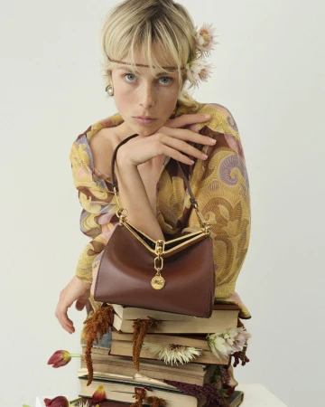 Model Edie Campbell holding an Etro Vela brown leather bag resting on a stack of books for the 2024 campaign.