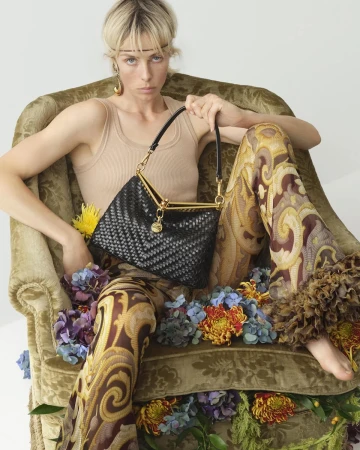 Model Edie Campbell holding a black woven leather bag while seated in a green upholstered armchair surrounded by flowers.