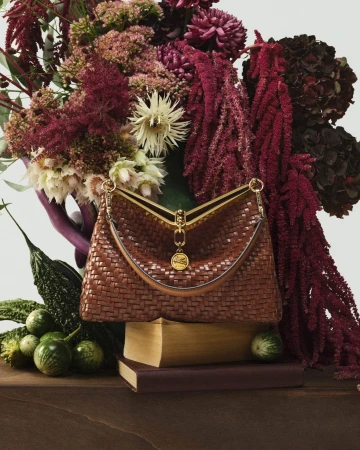 Brown woven leather handbag resting on stacked books before a lush arrangement of dark red flowers and vegetables