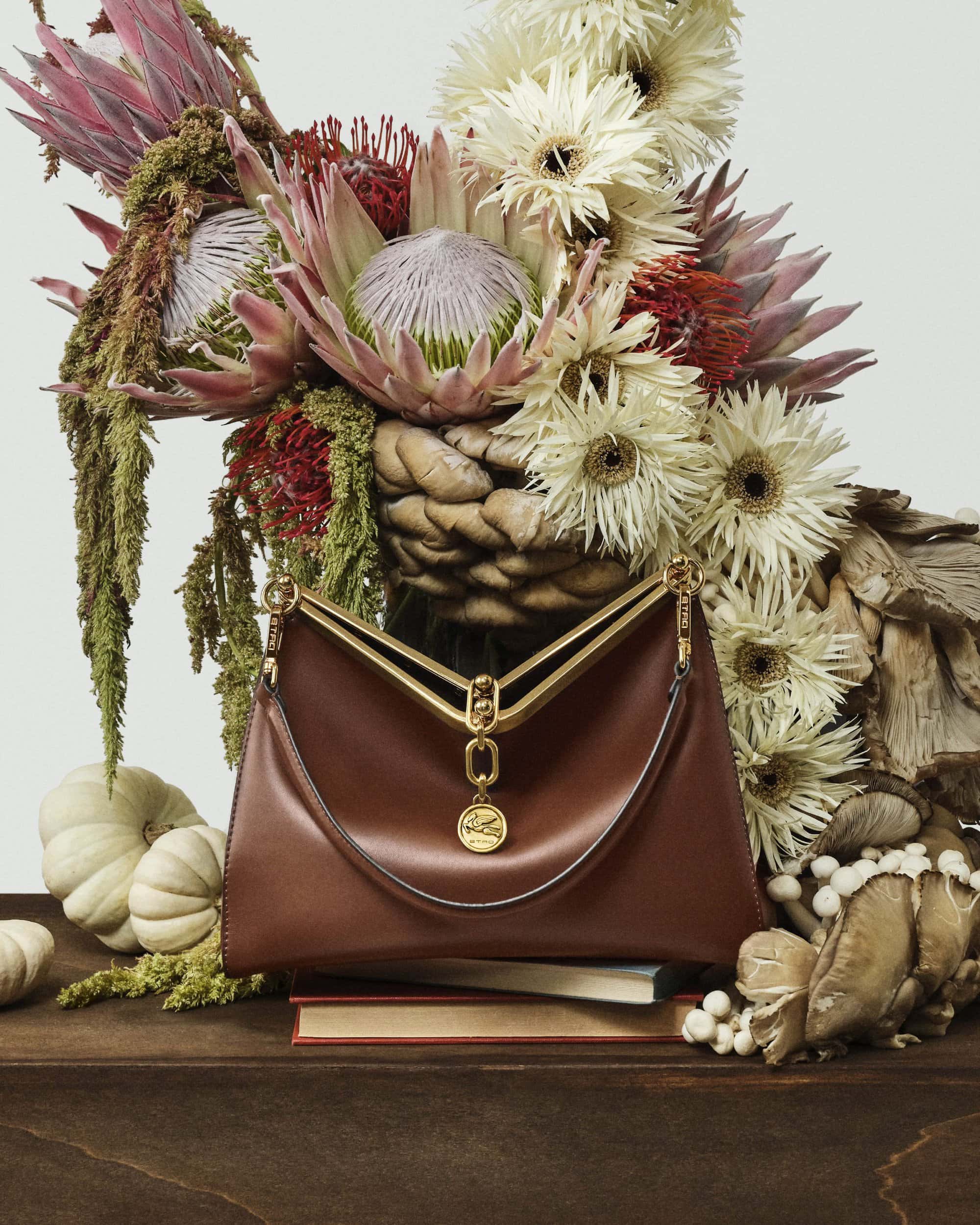 Brown leather handbag with gold hardware displayed in front of a large floral arrangement and white pumpkins on a dark wooden surface