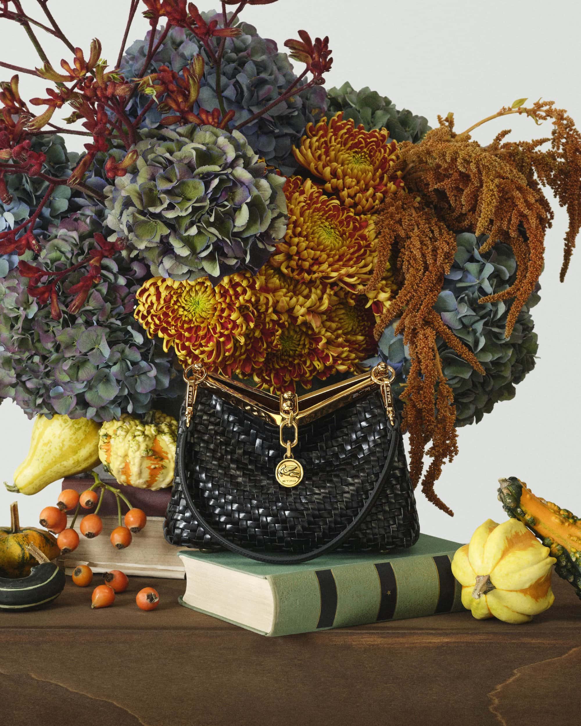 Black woven leather handbag resting on a green book before a large arrangement of autumn flowers and gourds.