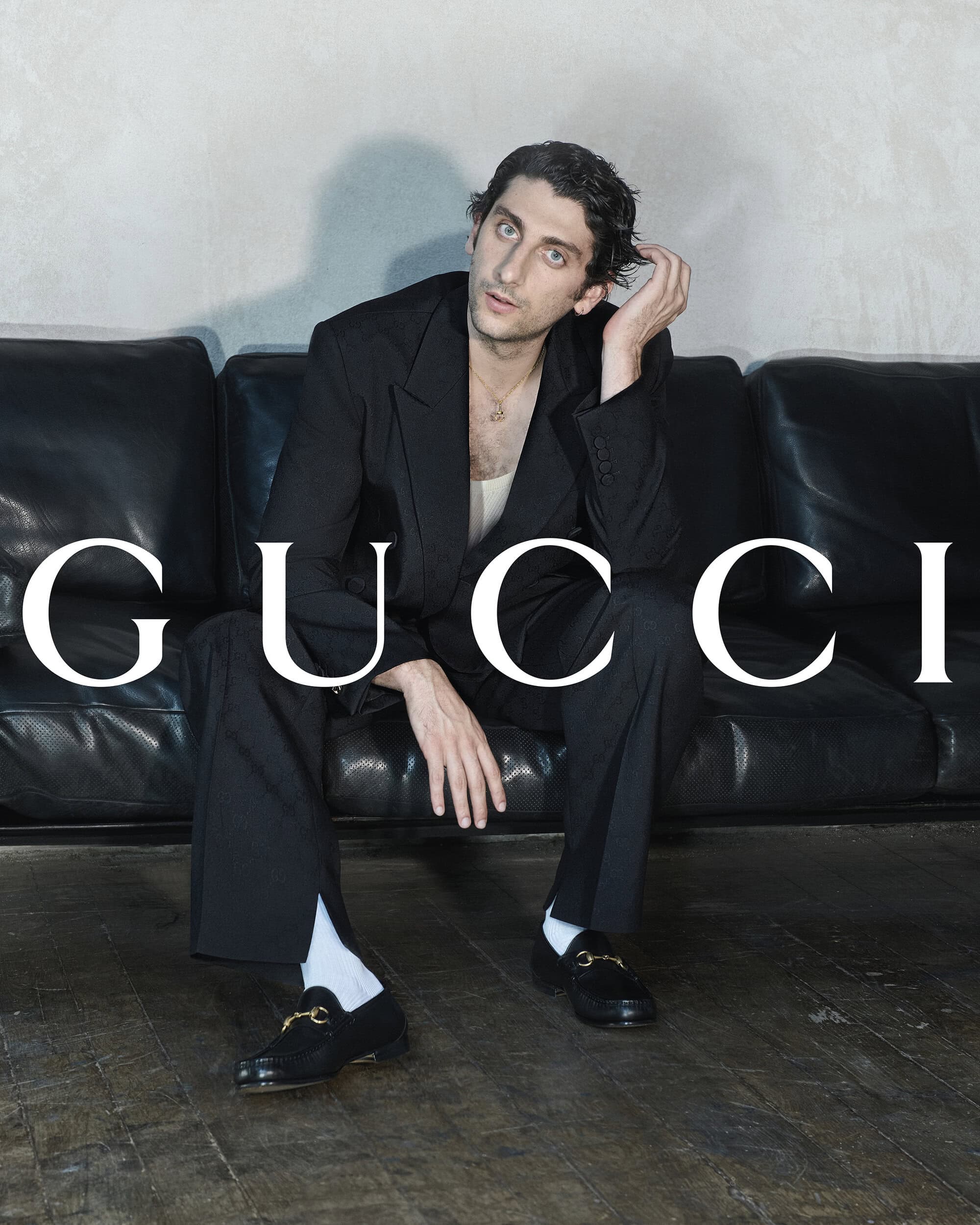 Pietro Castellitto seated on a black leather sofa wearing a black suit and horsebit loafers for Gucci