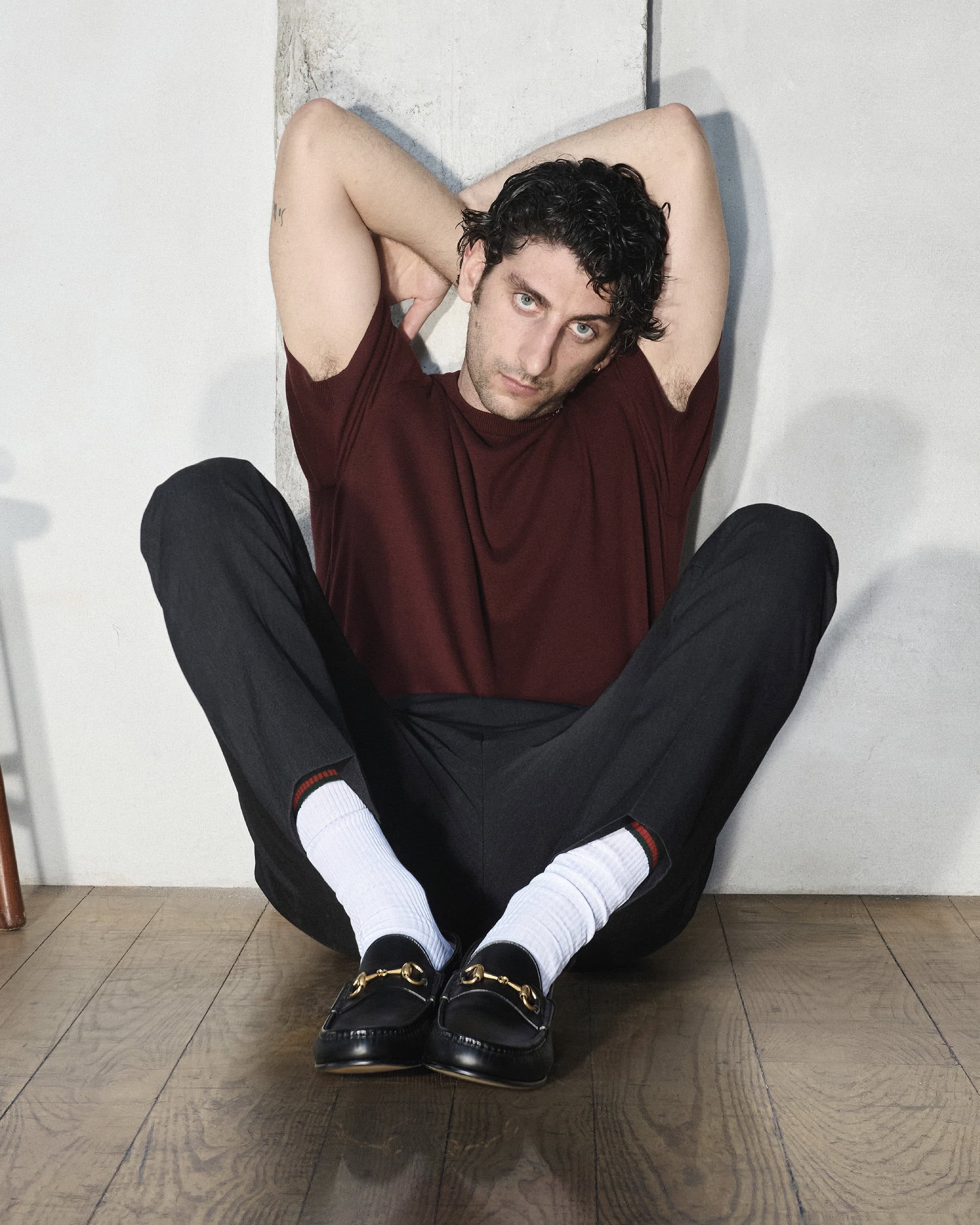 Model Pietro Castellitto seated against a wall wearing a burgundy t-shirt, dark trousers, white socks, and black leather loafers.