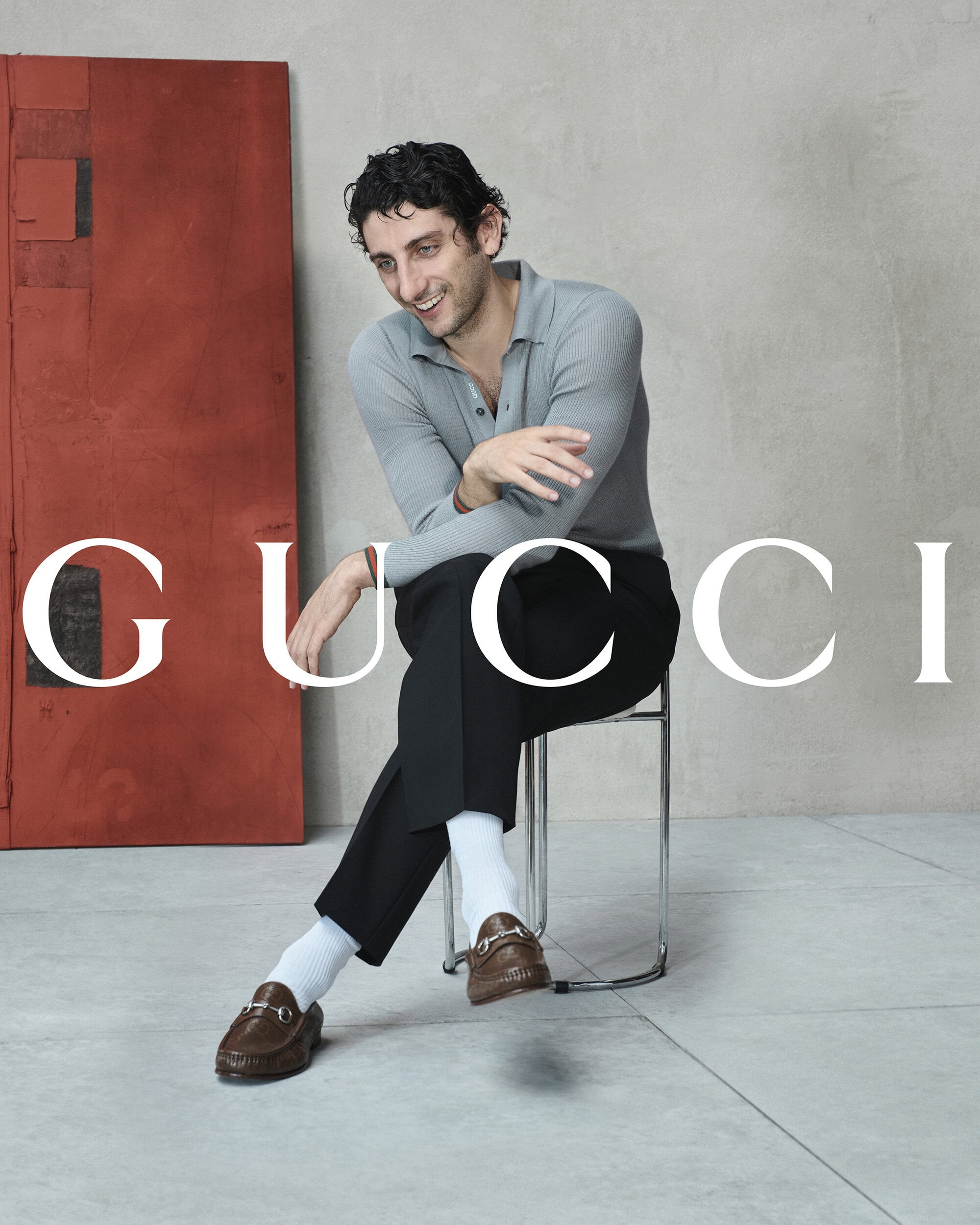 Pietro Castellitto seated on a metal chair wearing grey polo shirt and brown loafers for Gucci The Loafer Since 1953 ad campaign.