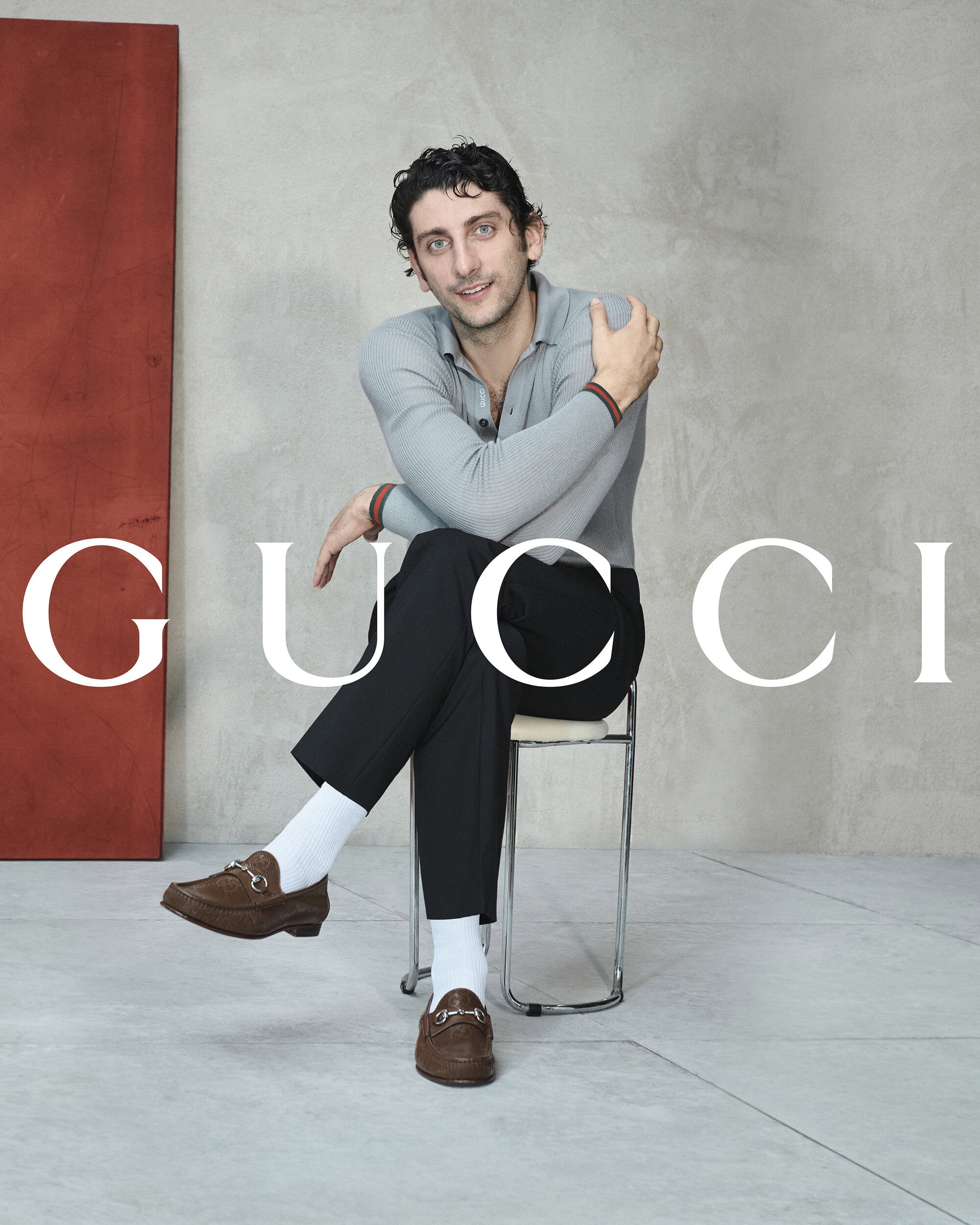 Pietro Castellitto seated on a wire chair against a concrete wall wearing a grey polo shirt, black trousers, white socks, and brown loafers.