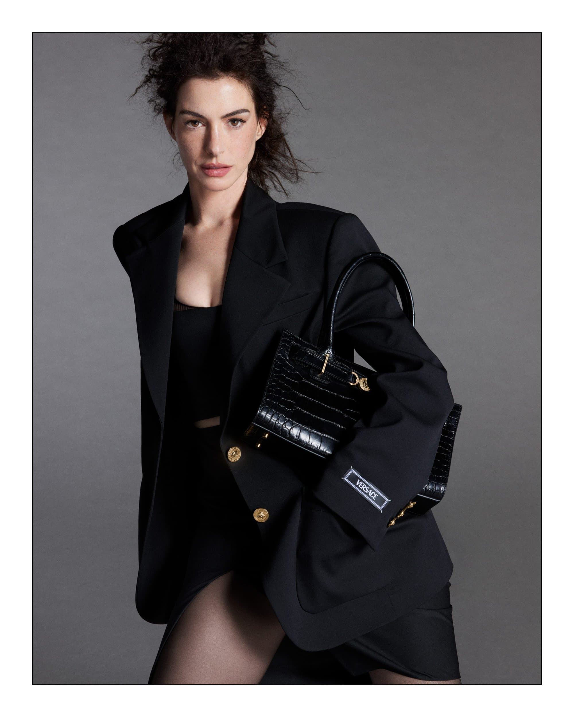 Anne Hathaway wearing a black blazer holding a black handbag against a grey studio background