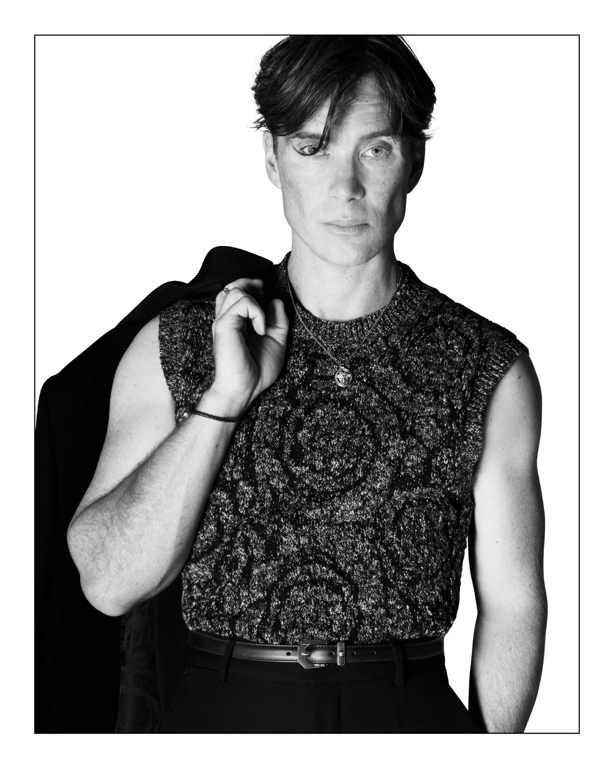 Monochrome portrait of Cillian Murphy wearing a patterned sleeveless knit top with a jacket draped over his shoulder against a white background