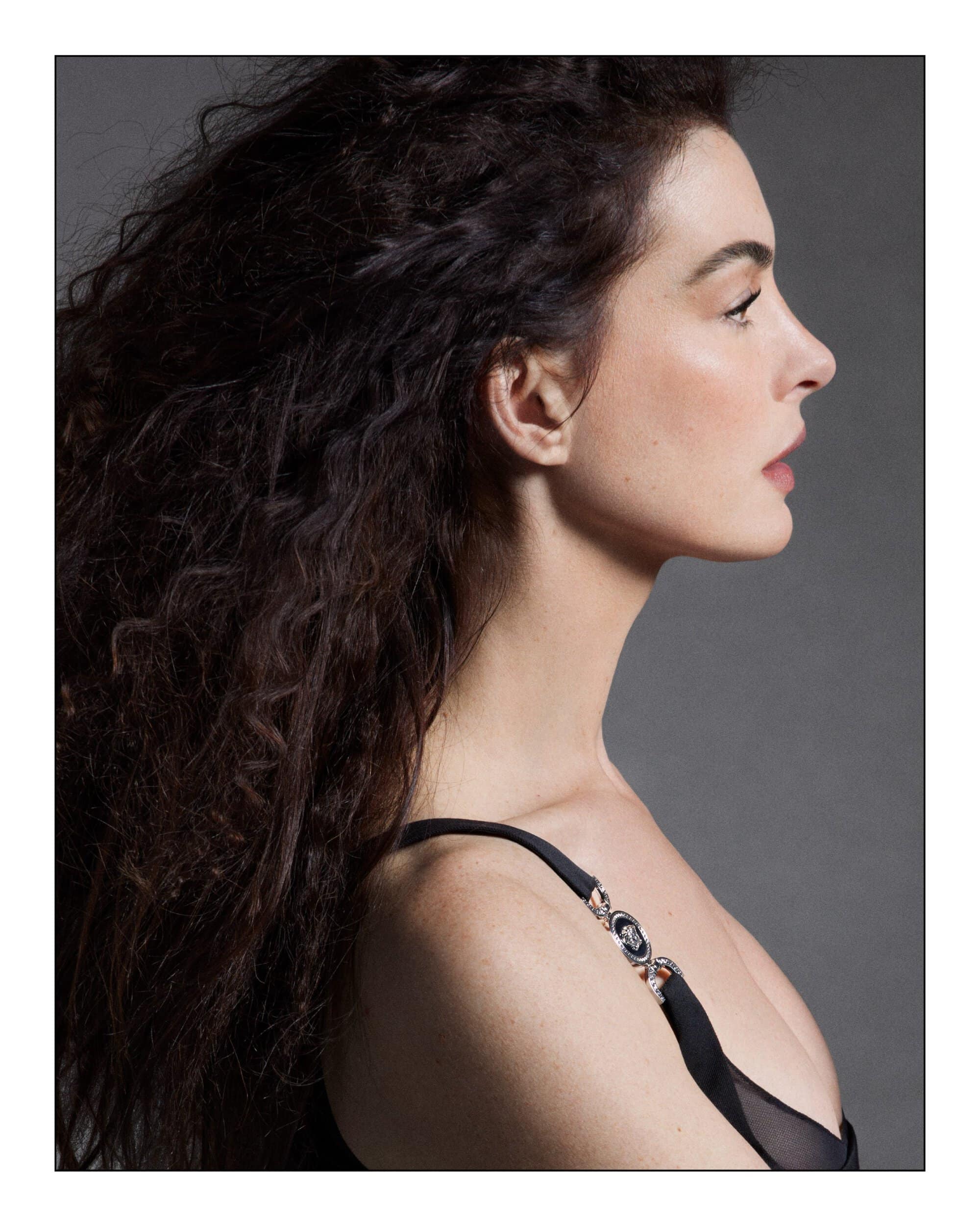 Profile portrait of actress Anne Hathaway with long dark curly hair wearing a black dress for Versace