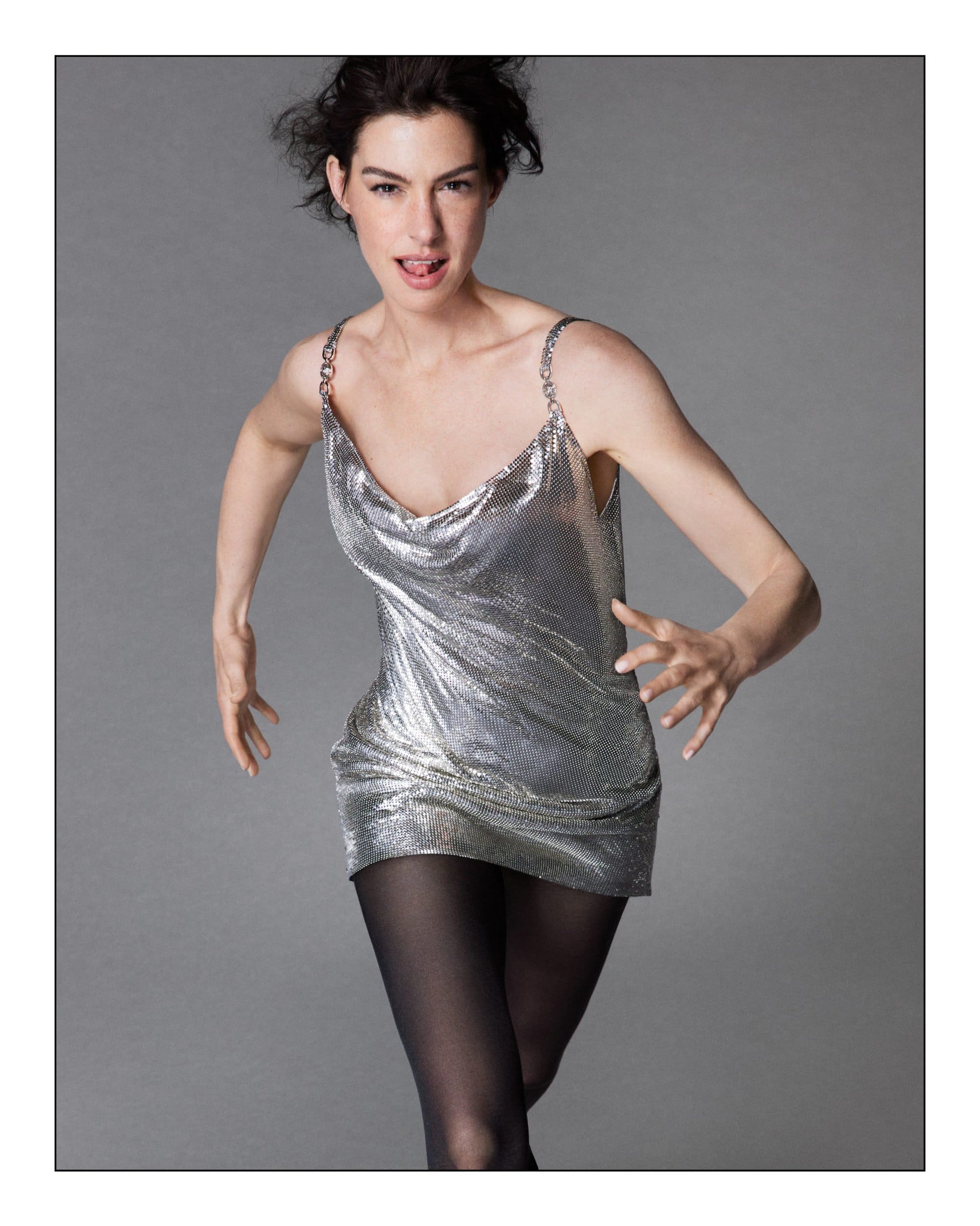Anne Hathaway wearing a metallic silver chainmail slip dress and dark tights against a plain grey studio background.