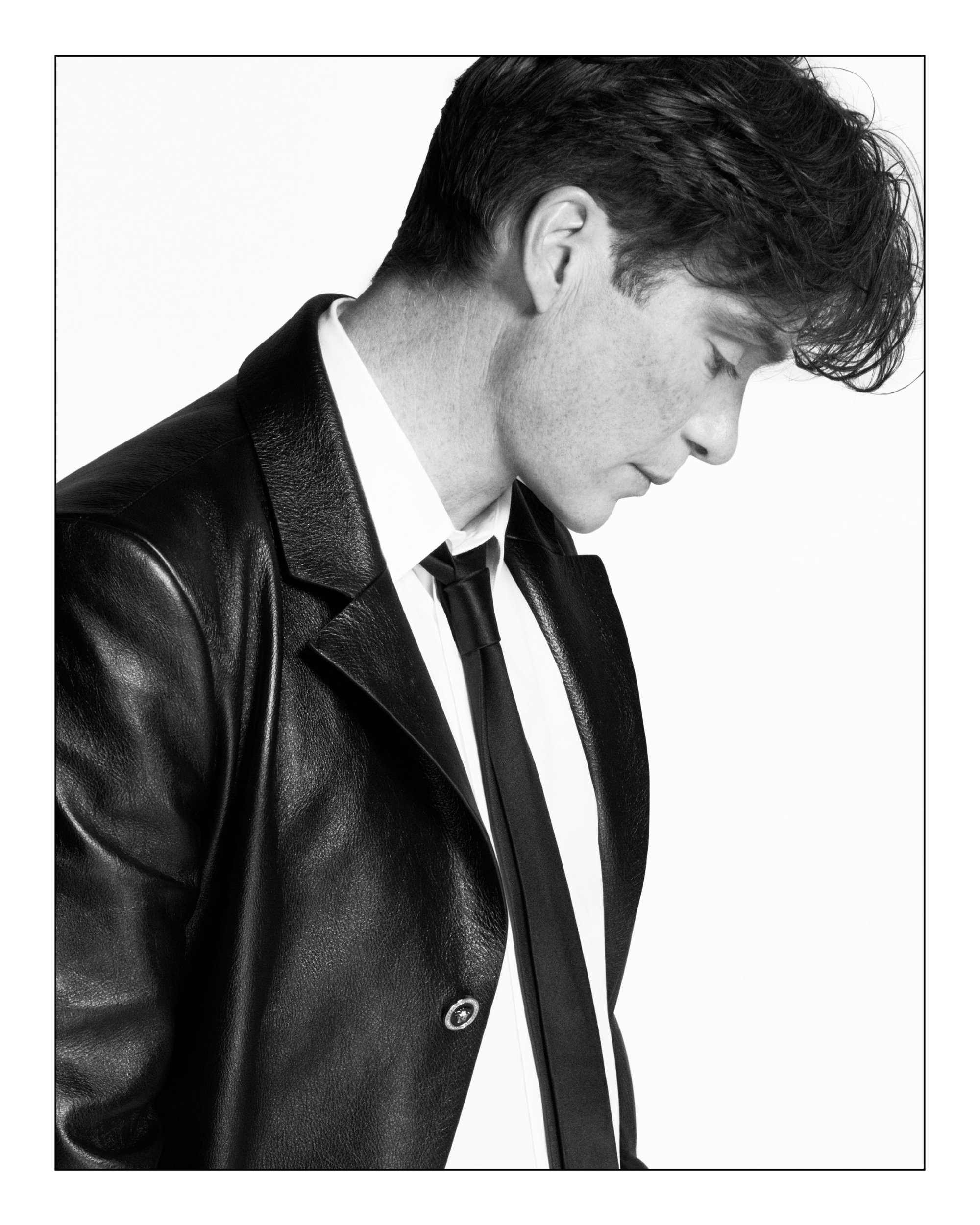 Monochrome portrait of Cillian Murphy in a black leather jacket and tie against a plain white background for Versace.