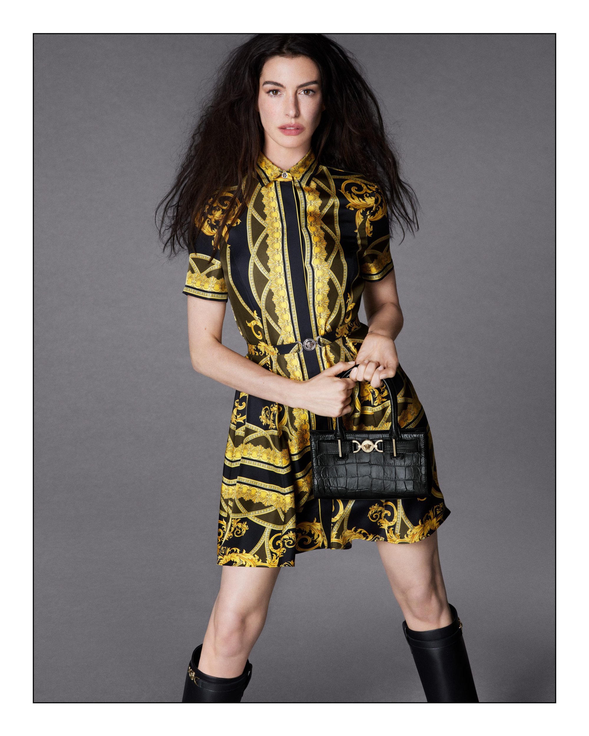 Anne Hathaway wearing a printed yellow and black dress holding a black handbag against a grey backdrop